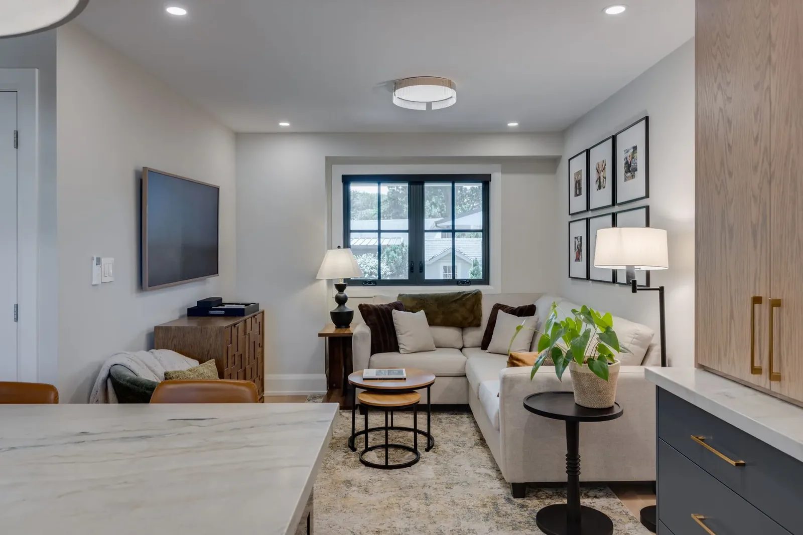 Family room open to the kitchen, with a putty sectional under a gallery wall of framed photographs, a wall-hung television over a carved wood cabinet and nesting tables on a patterned rug.