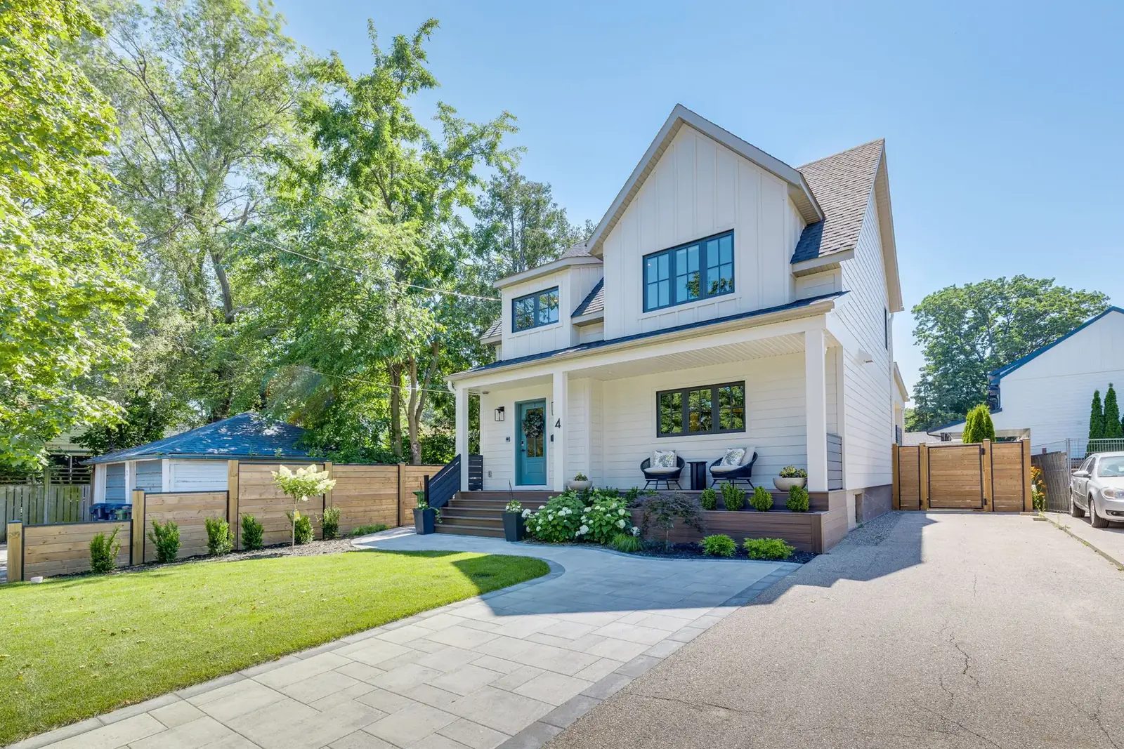 Front elevation of the finished two-storey home from an angle, board-and-batten gables over white siding, a covered porch with woven chairs and a wide paver walkway meeting the drive.