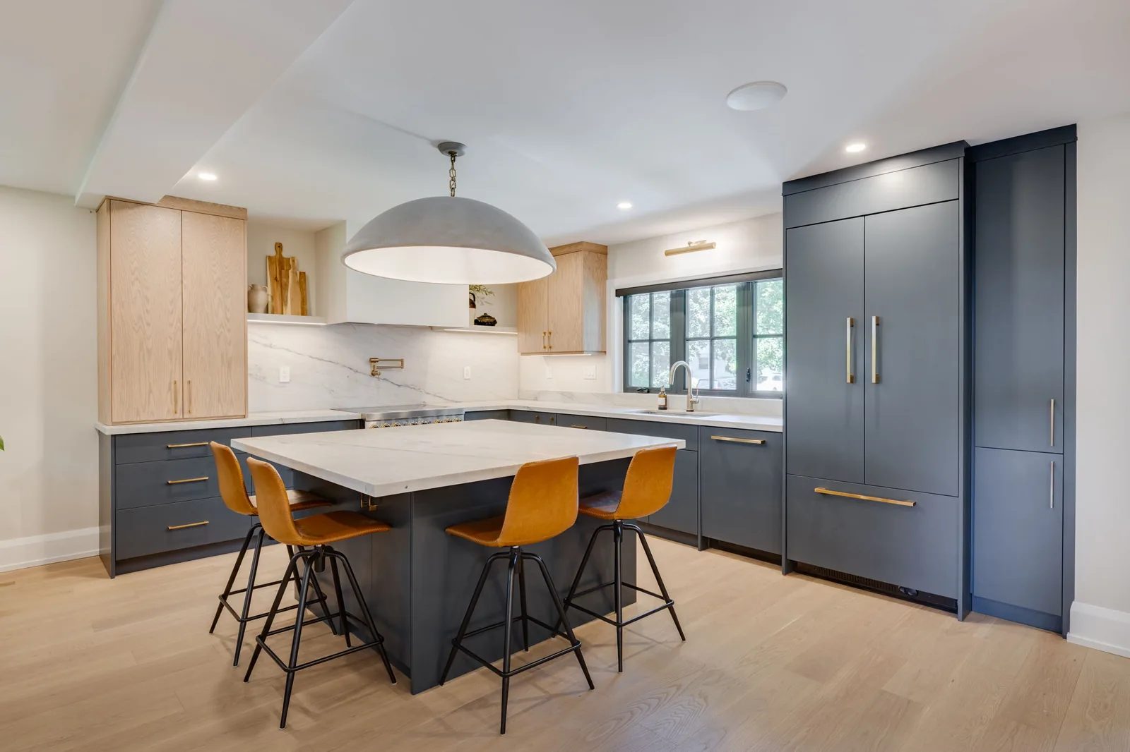 Open two-tone kitchen with white oak upper cabinets, navy base cabinets, a quartzite-topped island and amber leather counter stools under a dome pendant.