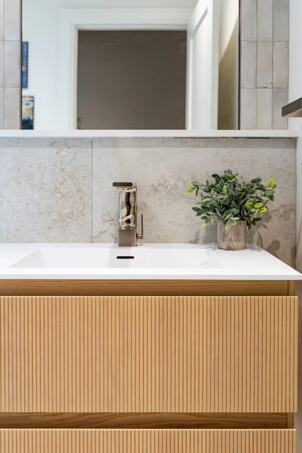 Fluted white oak drawer fronts beneath the integrated white sink and polished square faucet of the powder room, against stone-look tile.