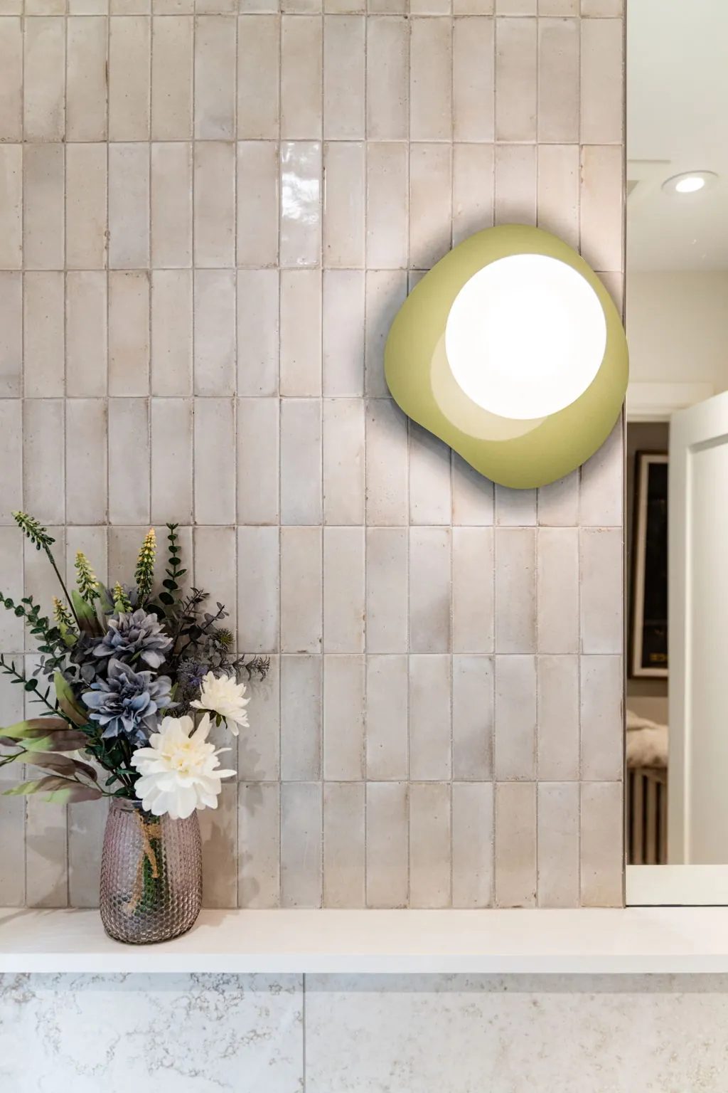 Sculptural olive wall sconce on the stacked vertical tile of the powder room, above a vase of flowers on the stone ledge.