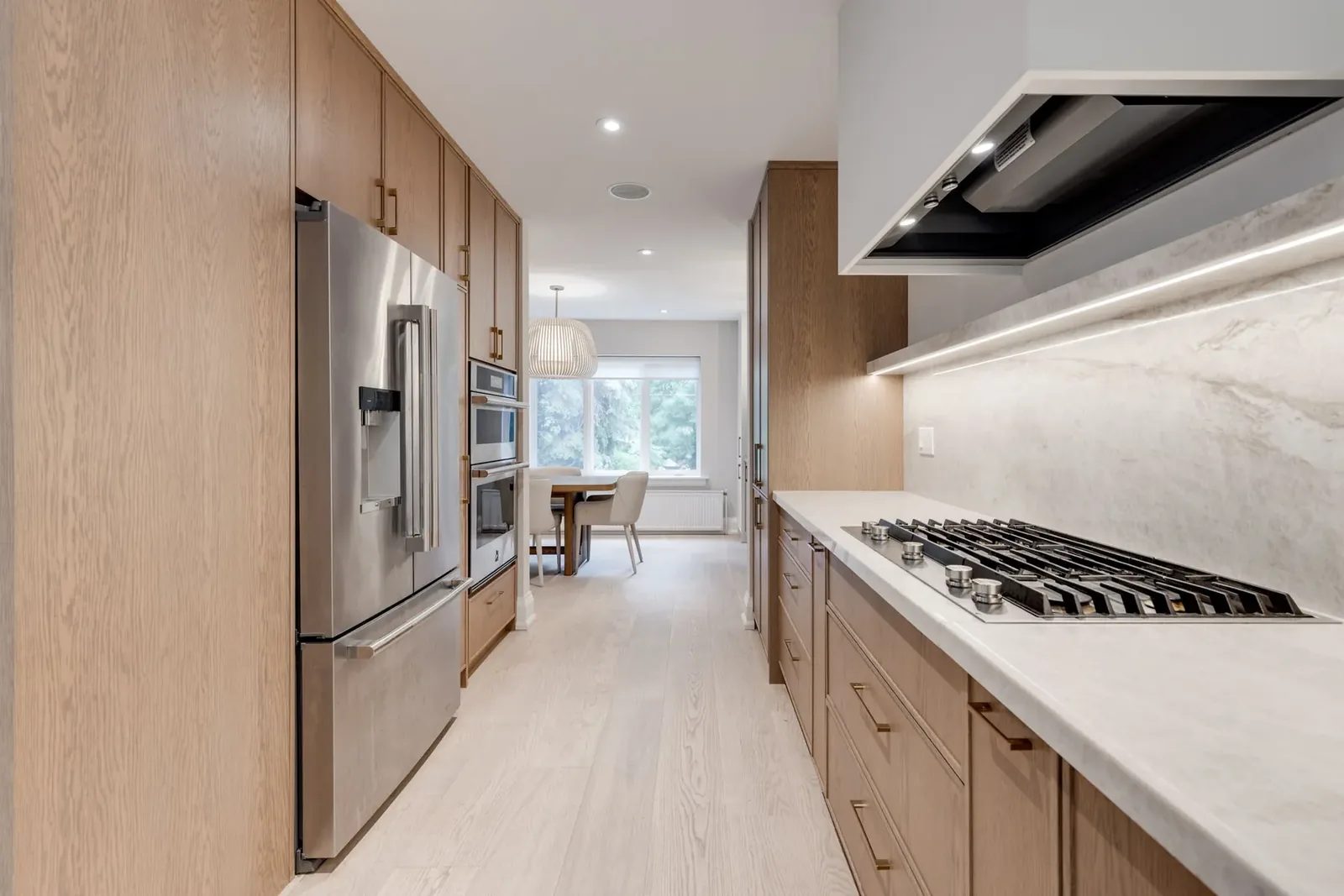 Down the galley kitchen, the stainless refrigerator and wall ovens facing the cooktop run, with the dining table beneath its slatted pendant beyond.