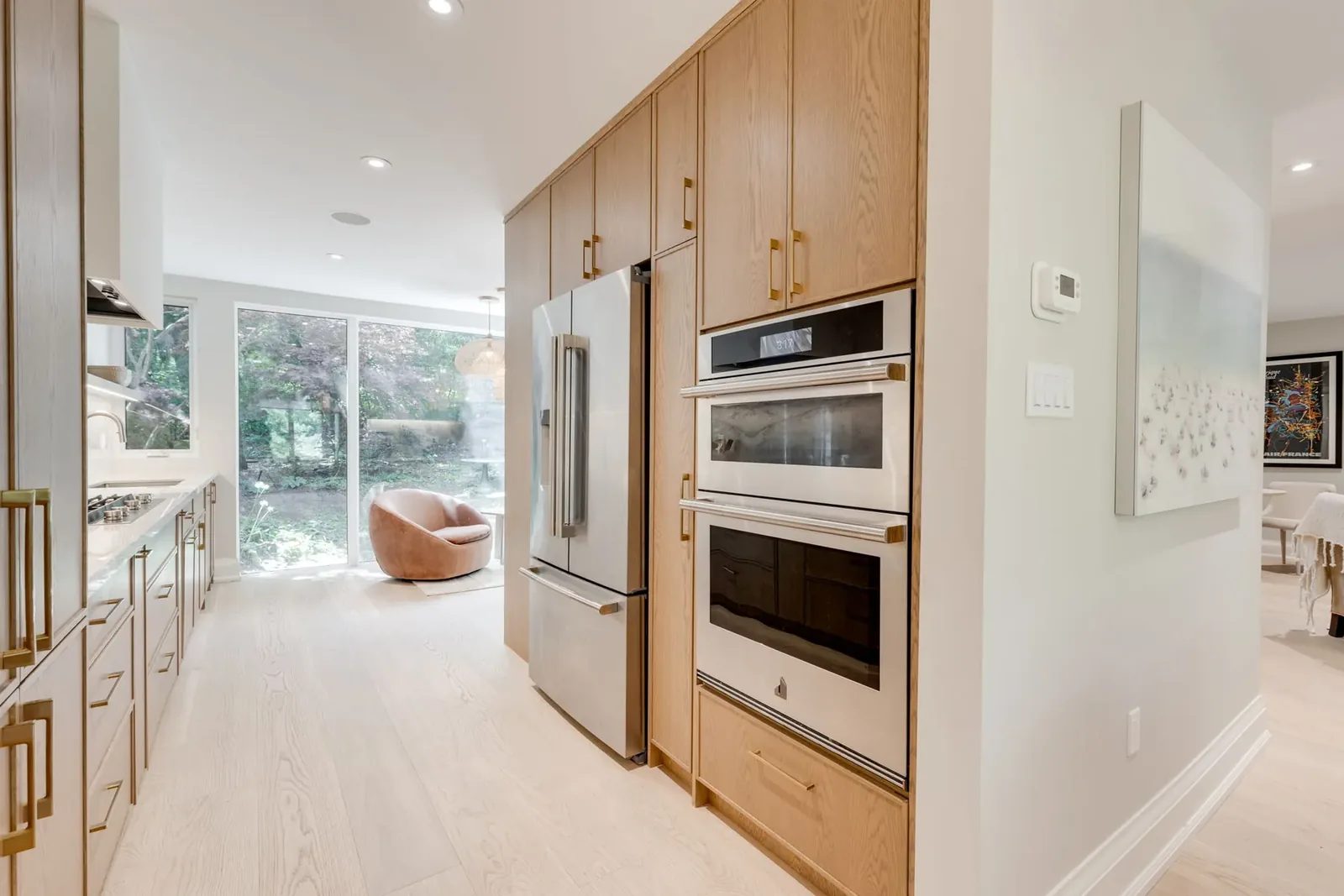 Tall white oak cabinetry holding the stainless refrigerator and integrated wall ovens, looking down the kitchen to the garden glass and a rust swivel chair.