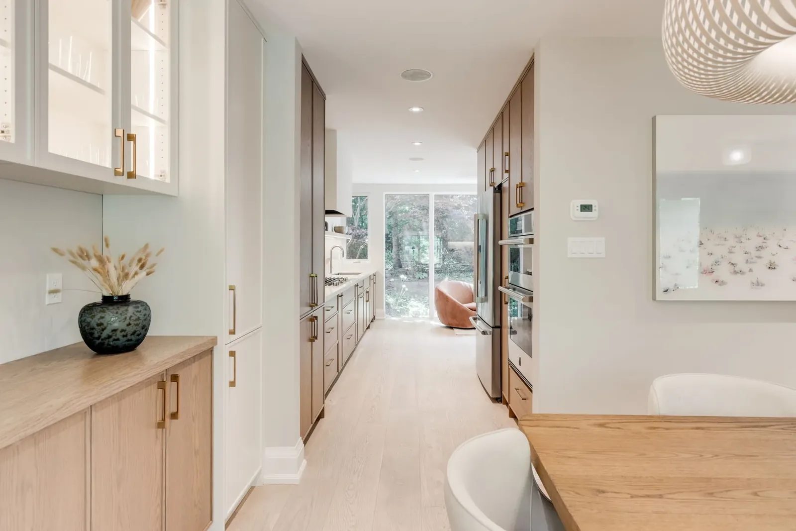 Galley kitchen seen from the servery, oak cabinetry and stone counters running to the full-height glass and a rust swivel chair at the garden end.