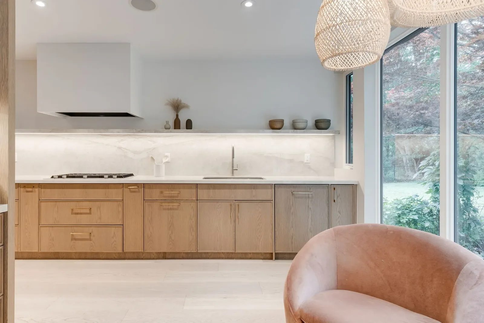 Kitchen run seen from the sitting nook, the plaster hood and lit stone shelf over the marble backsplash, with a blush swivel chair in the foreground.