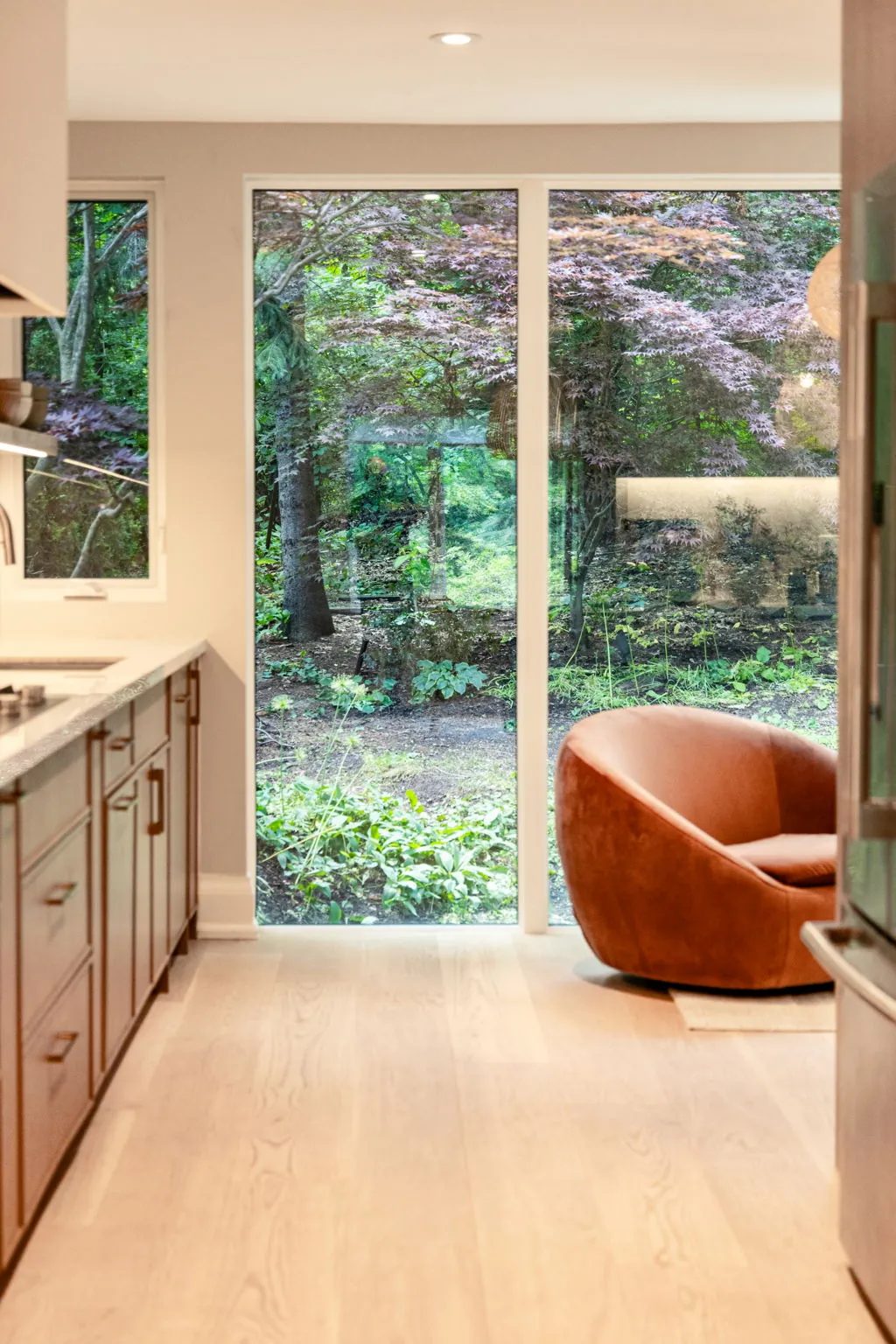 Floor-to-ceiling glass at the garden end of the kitchen, a rust leather swivel chair set against the view of the wooded yard.