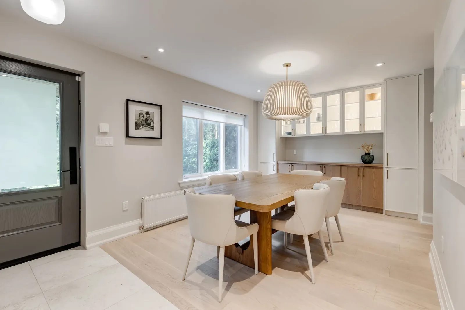 Dining room with an oak table and six cream upholstered chairs beneath a slatted wood pendant, backed by the glass-front servery wall.