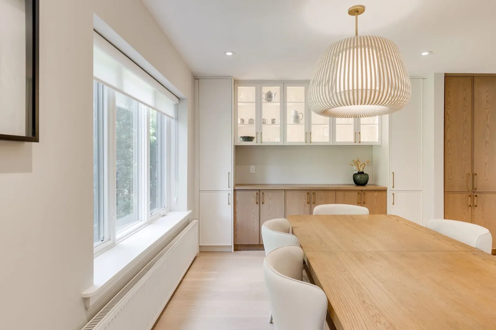 Dining room toward the servery, its lit glass-front uppers over white oak lower cabinets, with the slatted pendant over the table.