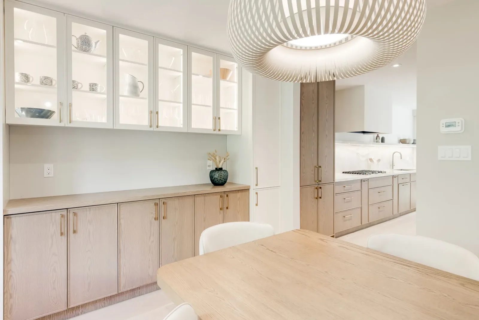 Dining servery with lit glass-front cabinets and oak lowers, looking through to the kitchen run and its marble backsplash.