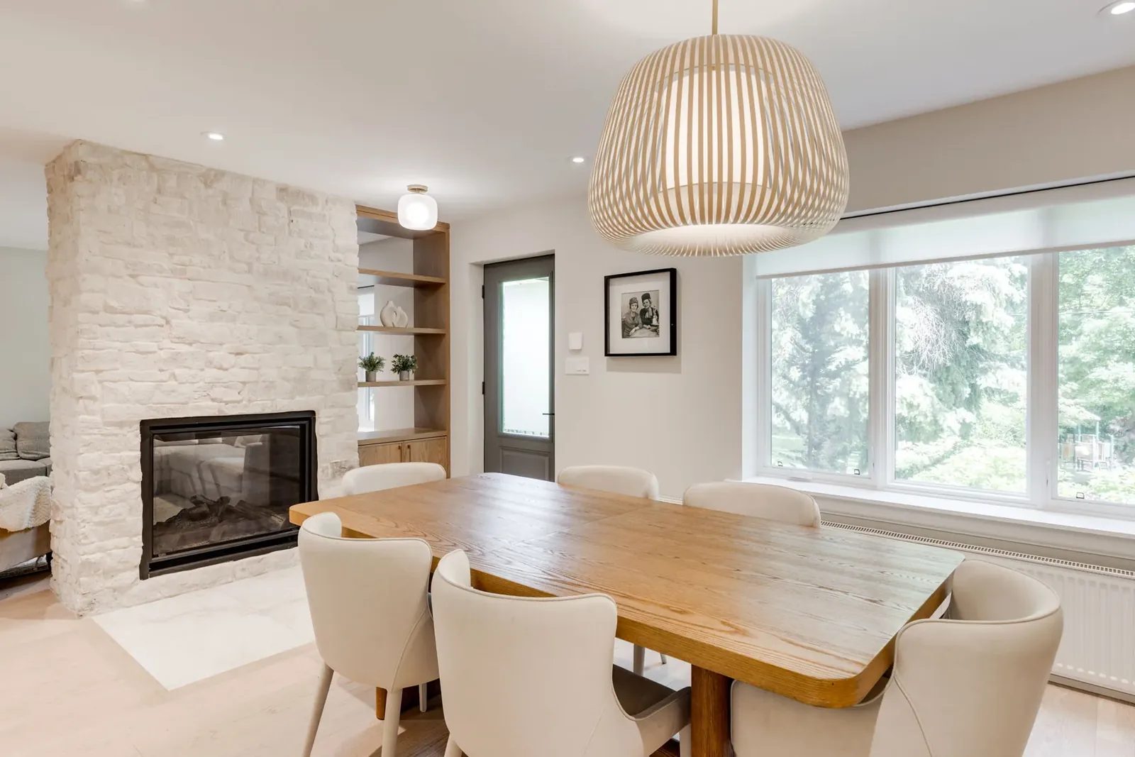 Dining table beneath the slatted wood pendant, set against the whitewashed stone fireplace and a picture window onto the trees.