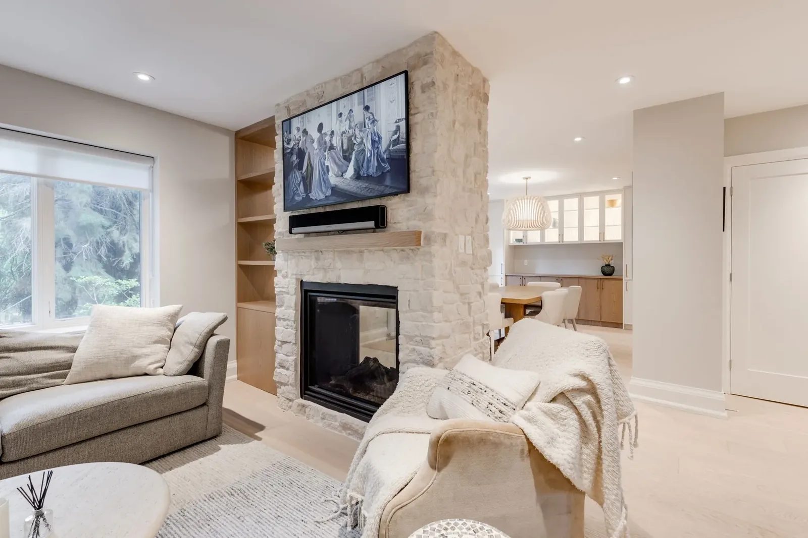 Living room at the two-sided whitewashed stone fireplace, a television and soundbar above the oak mantel, open to the dining room beyond.