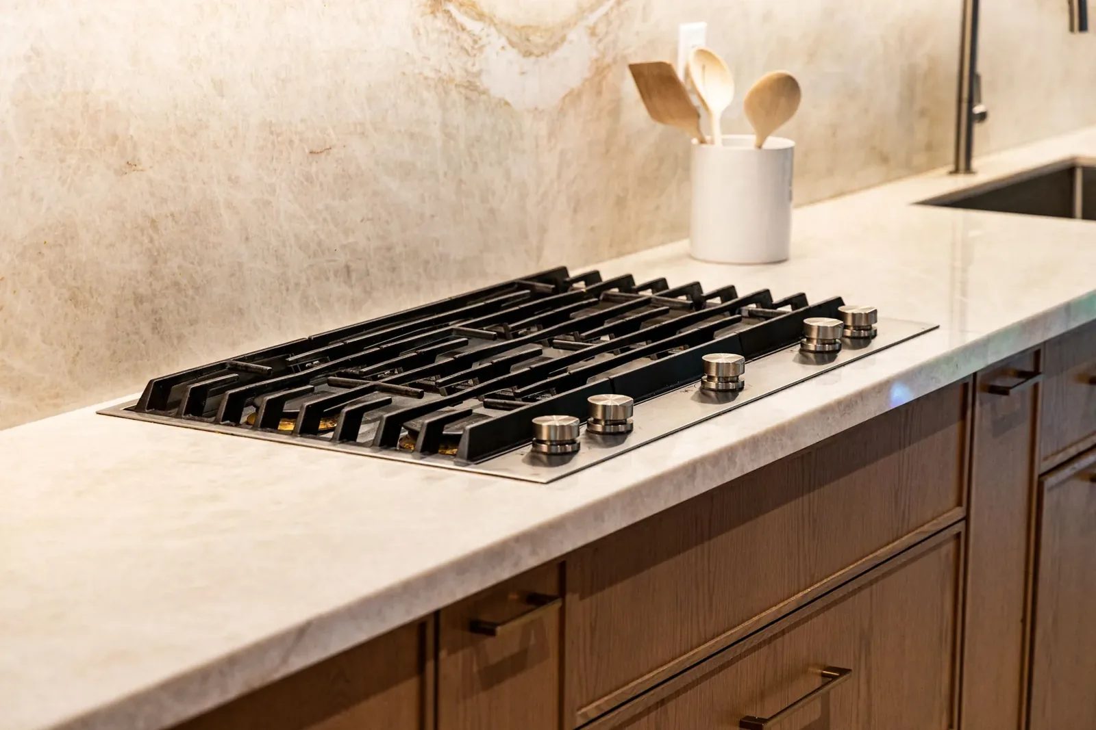 Gas cooktop with cast-iron grates set flush into the stone counter, a crock of wooden spoons against the marble slab backsplash and the sink beyond.