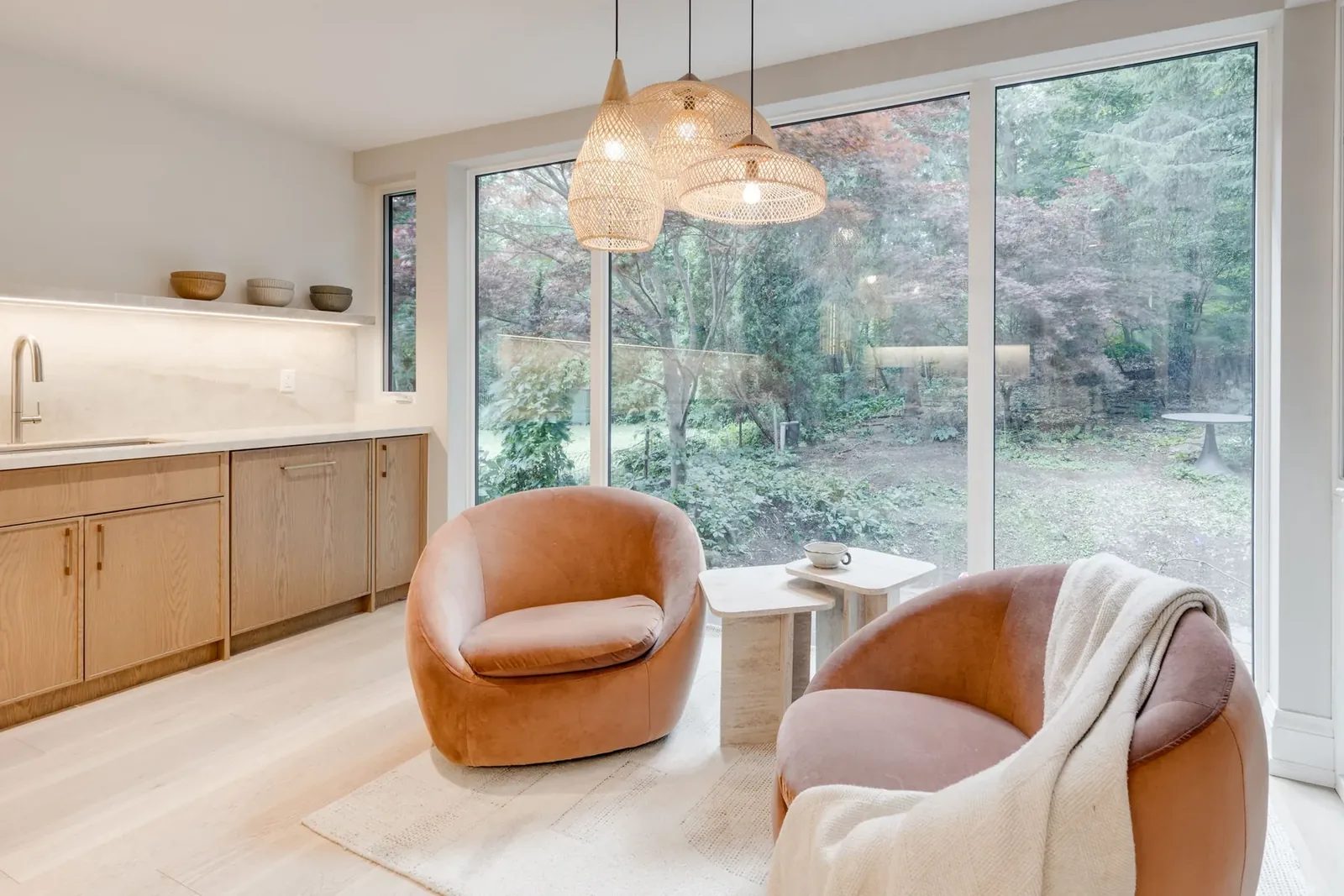 Sitting nook of two velvet swivel chairs and travertine side tables under a cluster of woven pendants, facing the wall of glass onto the wooded garden, with the servery counter alongside.