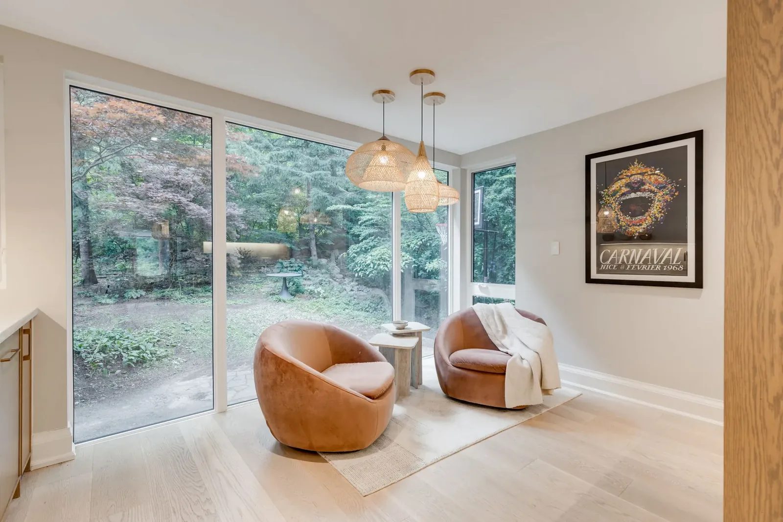 Sitting area with two velvet swivel chairs and a travertine side table on a pale rug, framed by full-height glass onto the ravine, a framed print on the end wall.
