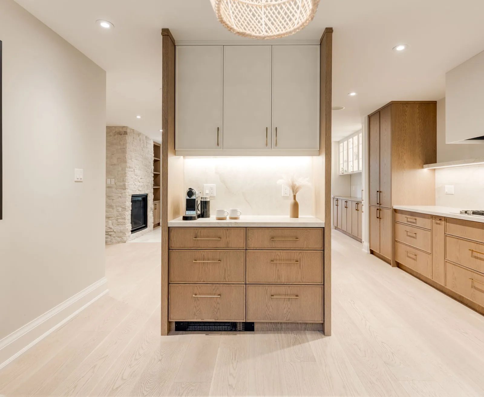 White oak servery peninsula with a coffee station and stone counter beneath a woven pendant, opening onto the kitchen run and the stone fireplace beyond.