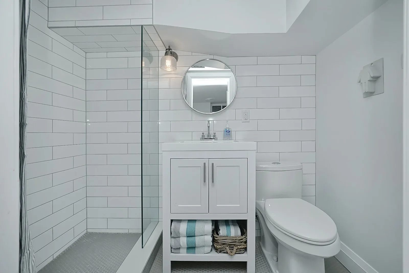 Bright renovated bathroom with white subway tile, glass shower, round mirror and compact vanity storage.