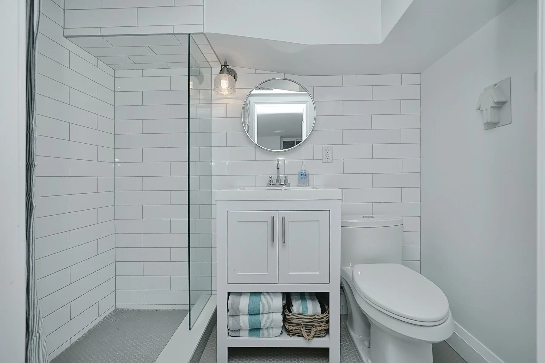 Bright renovated bathroom with white subway tile, glass shower, round mirror and compact vanity storage.