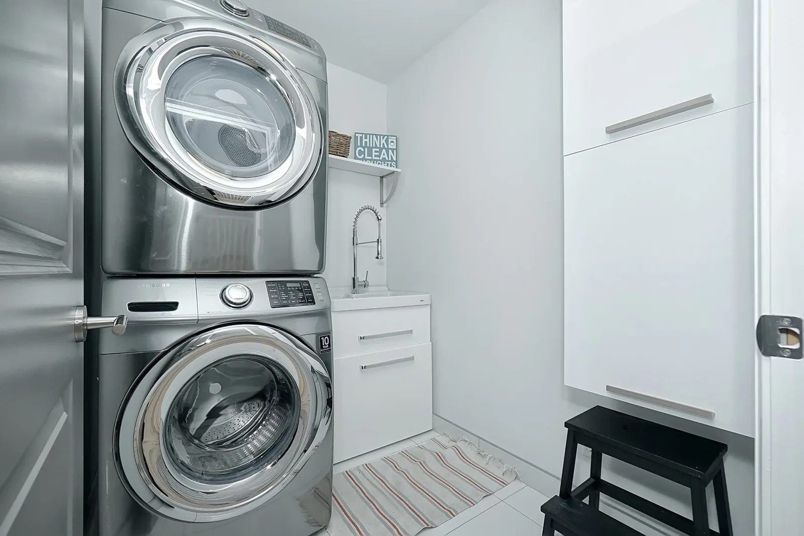 Bright white laundry room with stacked stainless appliances, organised cabinets, sink, and tiled floor.
