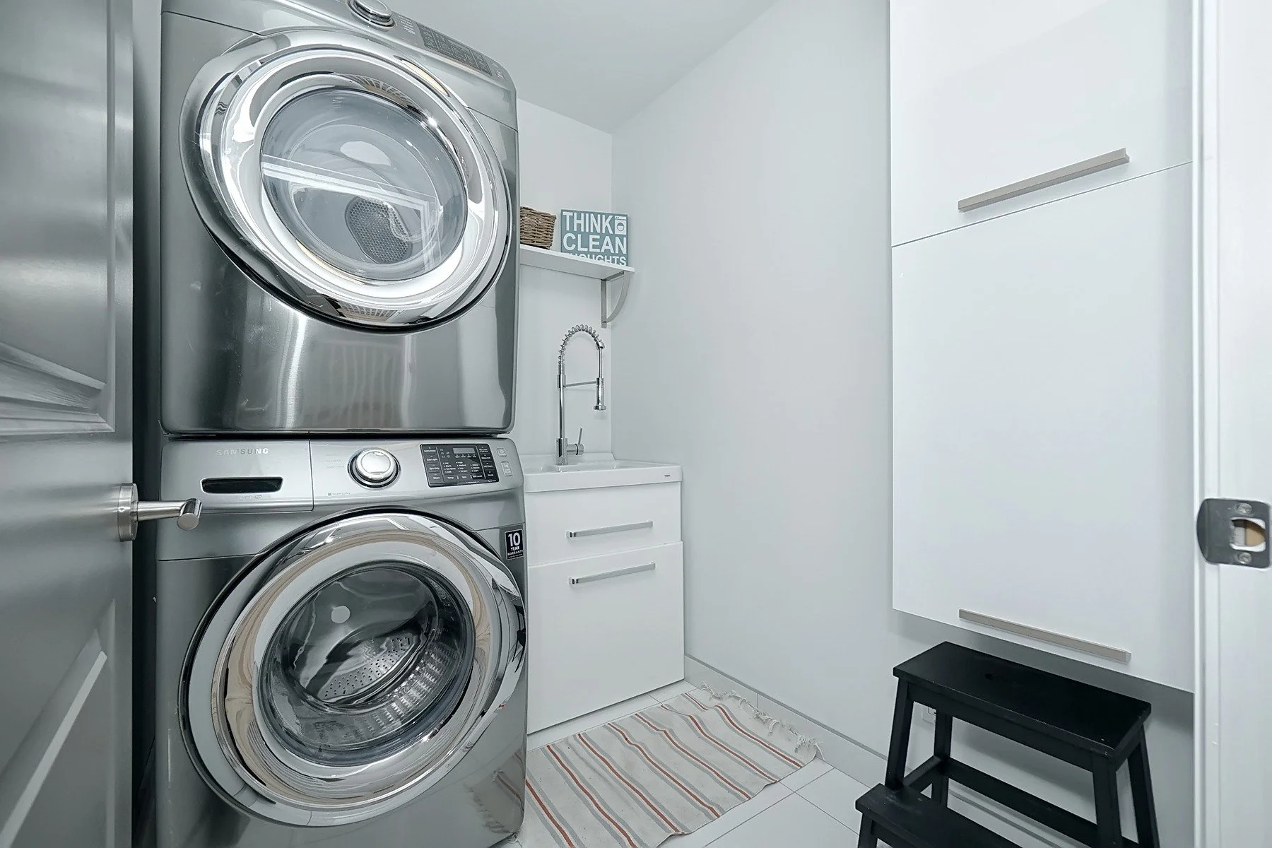 Bright white laundry room with stacked stainless appliances, organised cabinets, sink, and tiled floor.