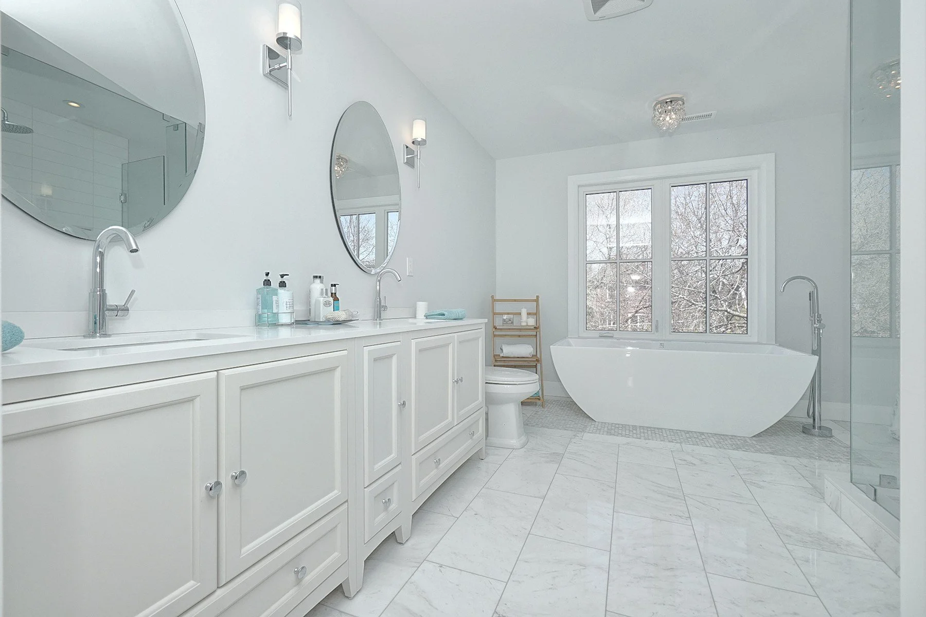 Bright white bathroom renovation with a double vanity, freestanding tub, glass shower, and marble tile floor.