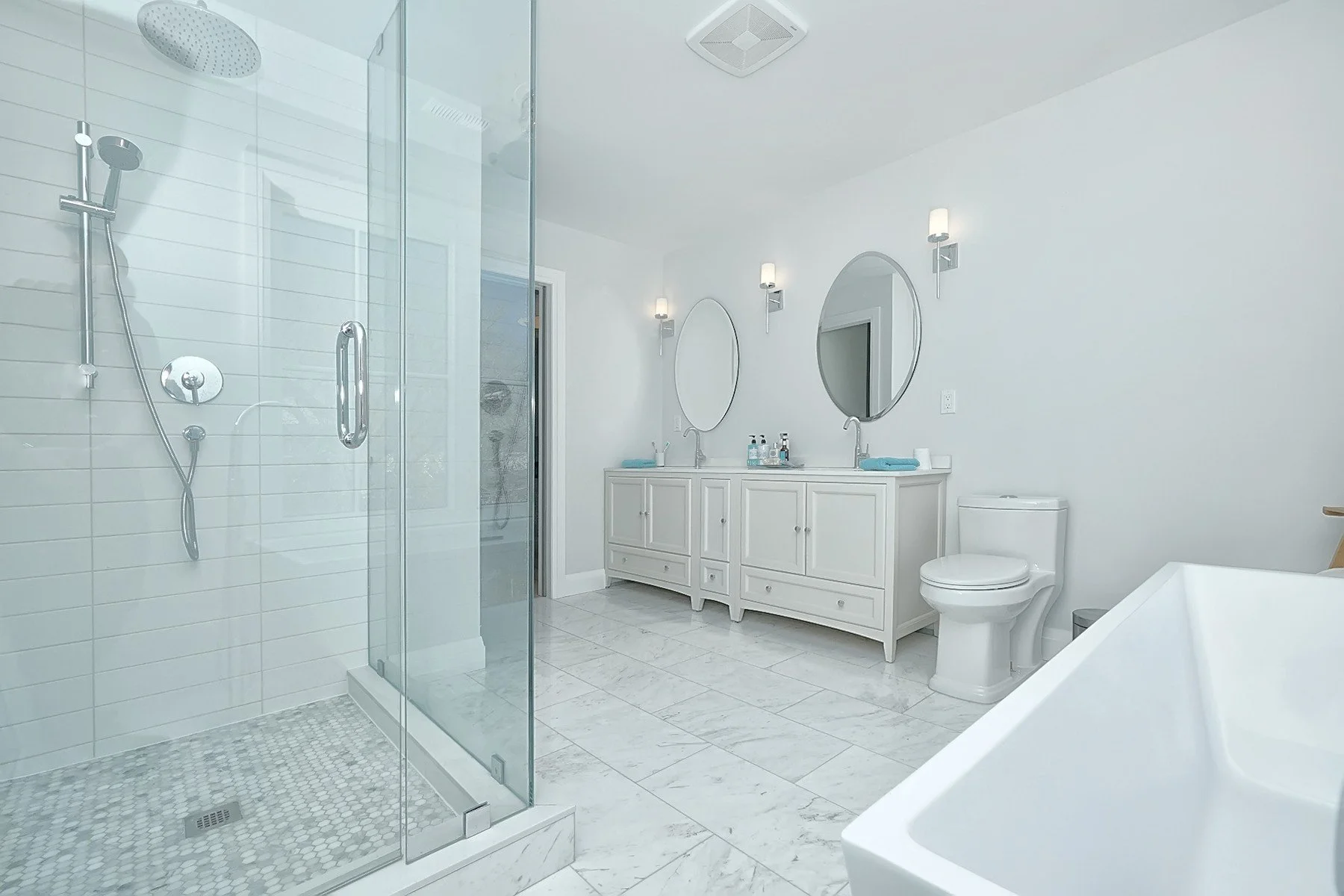 Bright white renovated bathroom with glass shower, double vanity, round mirrors and freestanding tub.