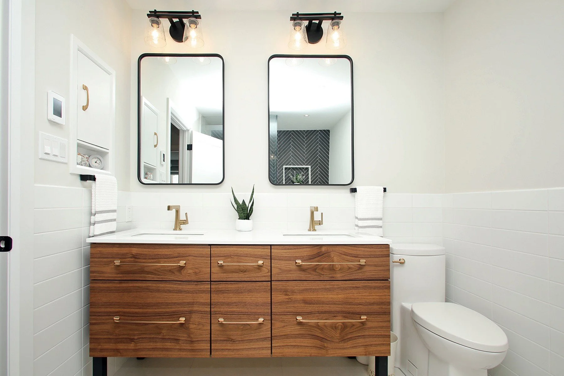 Bright bathroom with a walnut vanity, black mirrors, brass fixtures, and white tile walls.