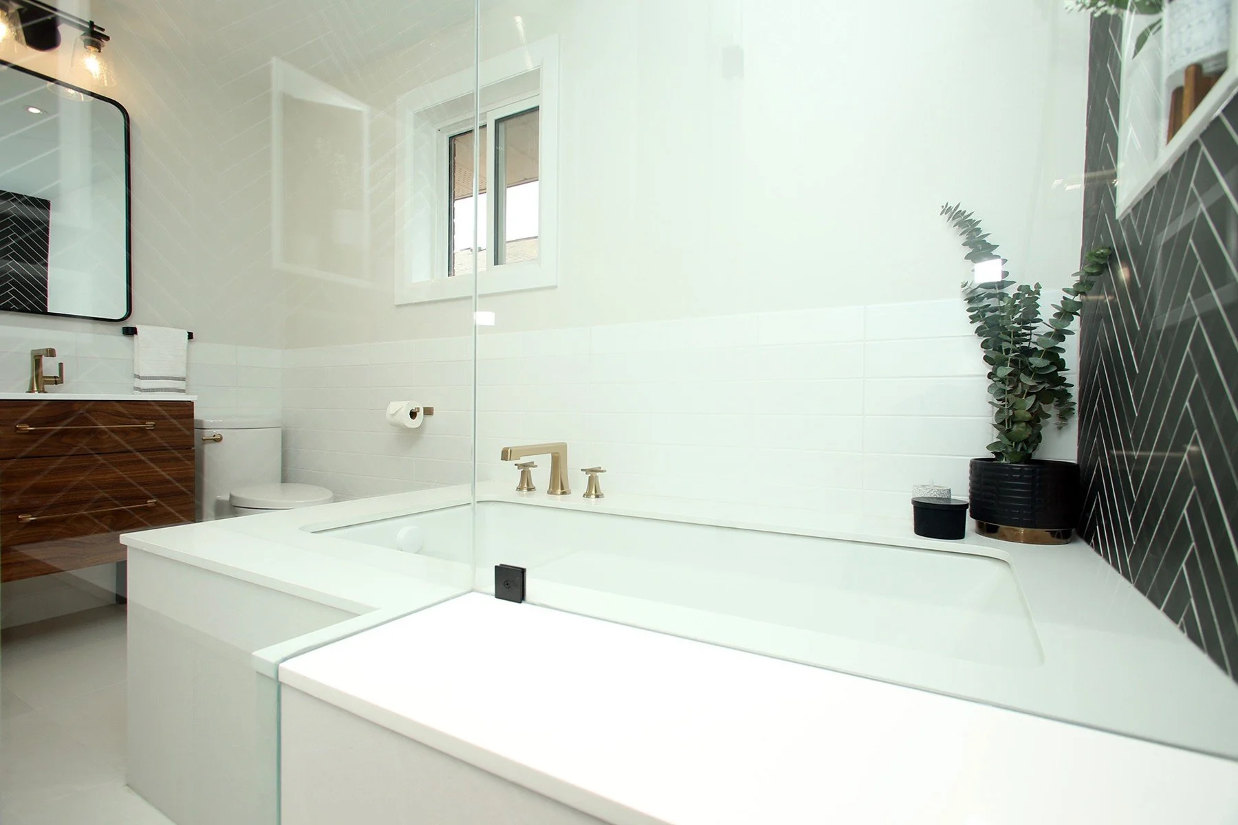 Bright white bathroom with a built-in tub, brass fixtures, dark herringbone tile and a wood vanity.