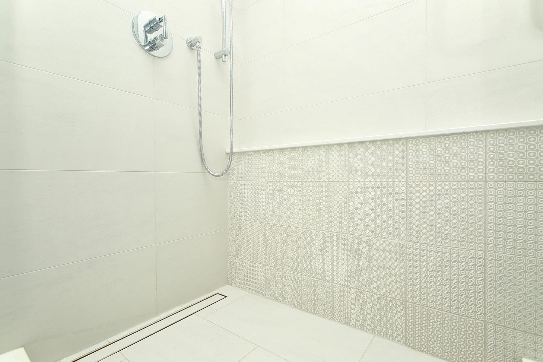Warm white walk-in shower with patterned accent tile, chrome handheld fixture and a linear drain.