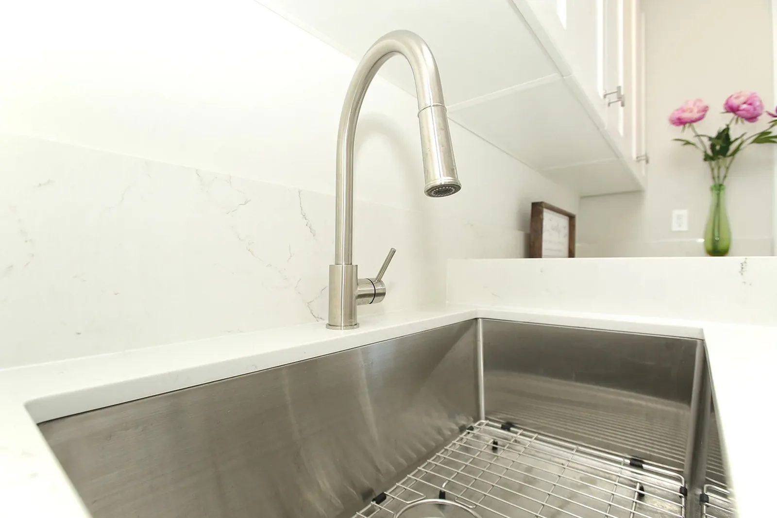 Large stainless sink and brushed metal faucet set into warm white quartz in a bright, organised kitchen.