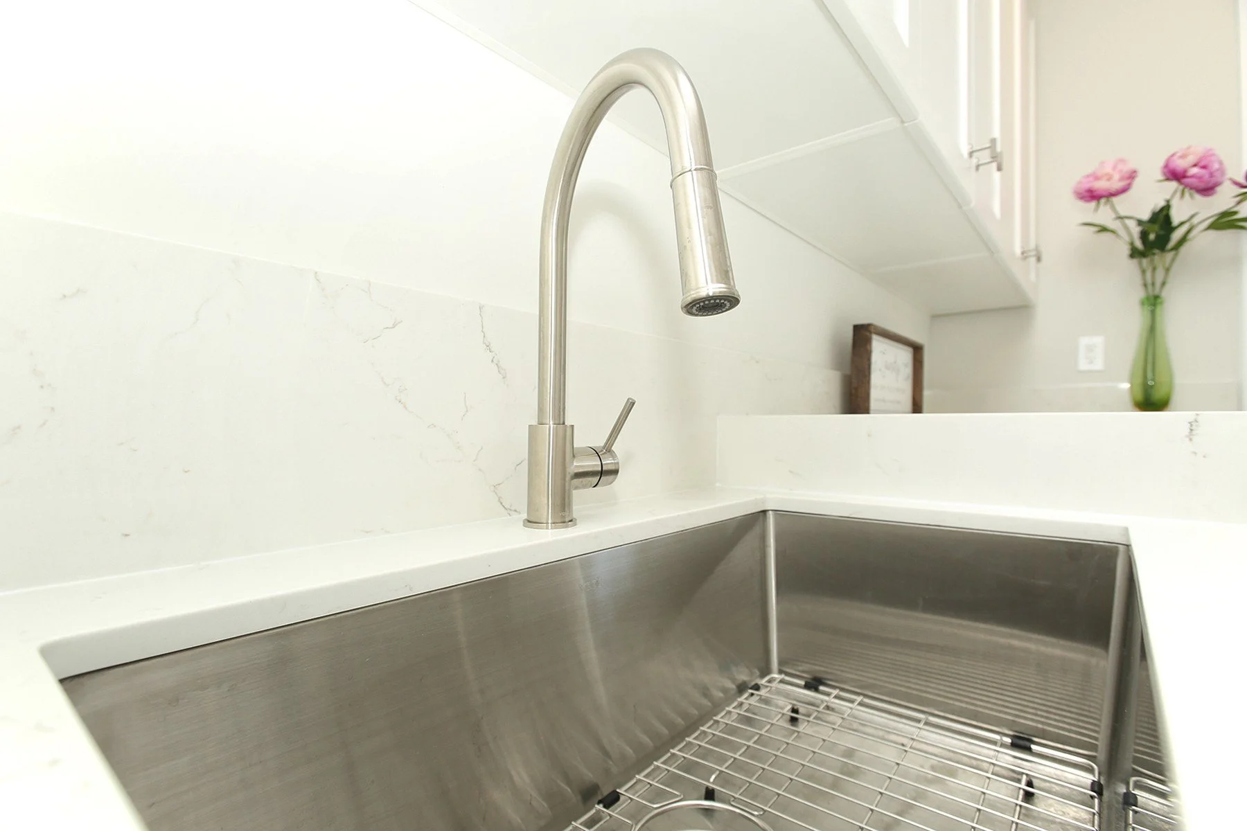 Large stainless sink and brushed metal faucet set into warm white quartz in a bright, organised kitchen.