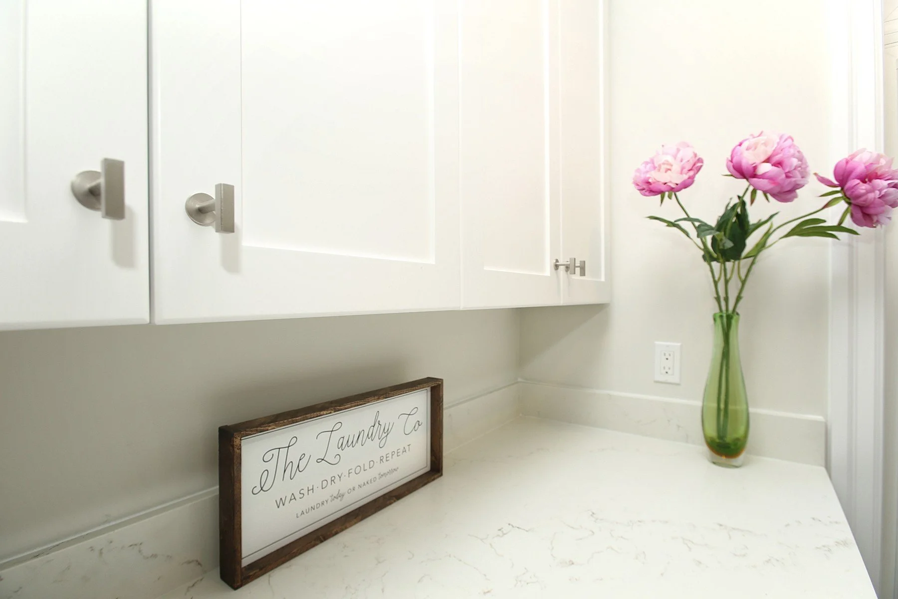 Warm white laundry room detail with shaker cabinets, quartz counter and pink peonies in a green vase.