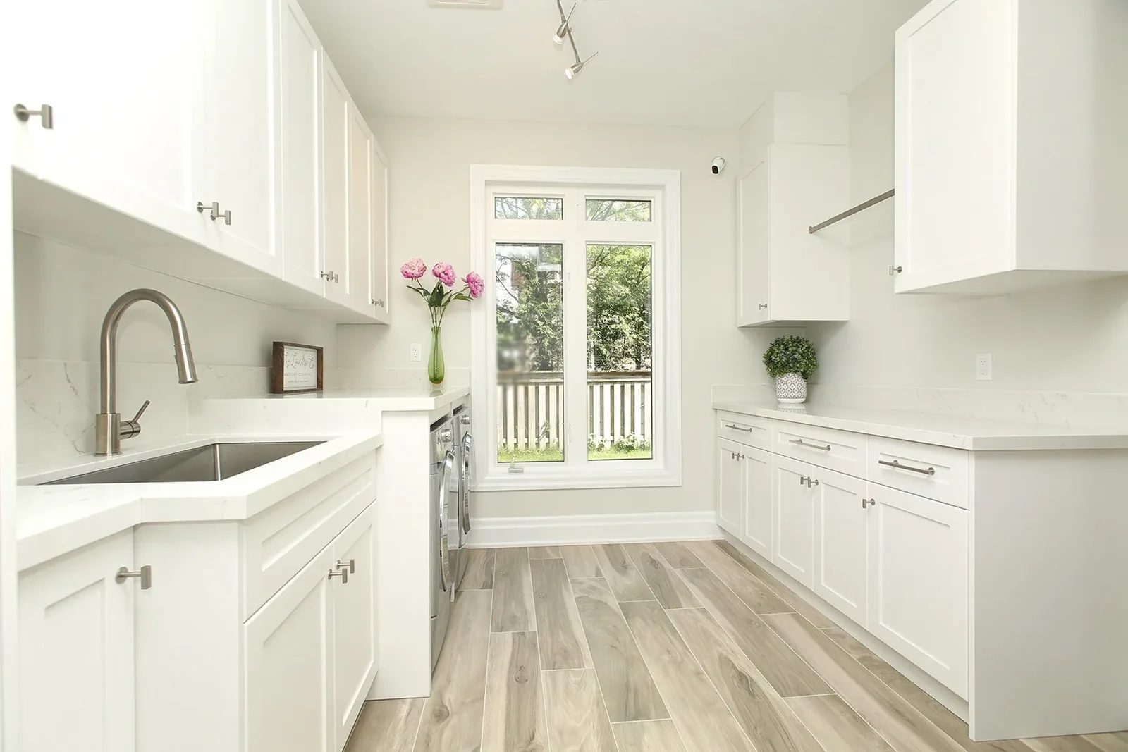 Bright laundry room with white shaker cabinets, quartz counters, grey floor tile and a tall rear window.