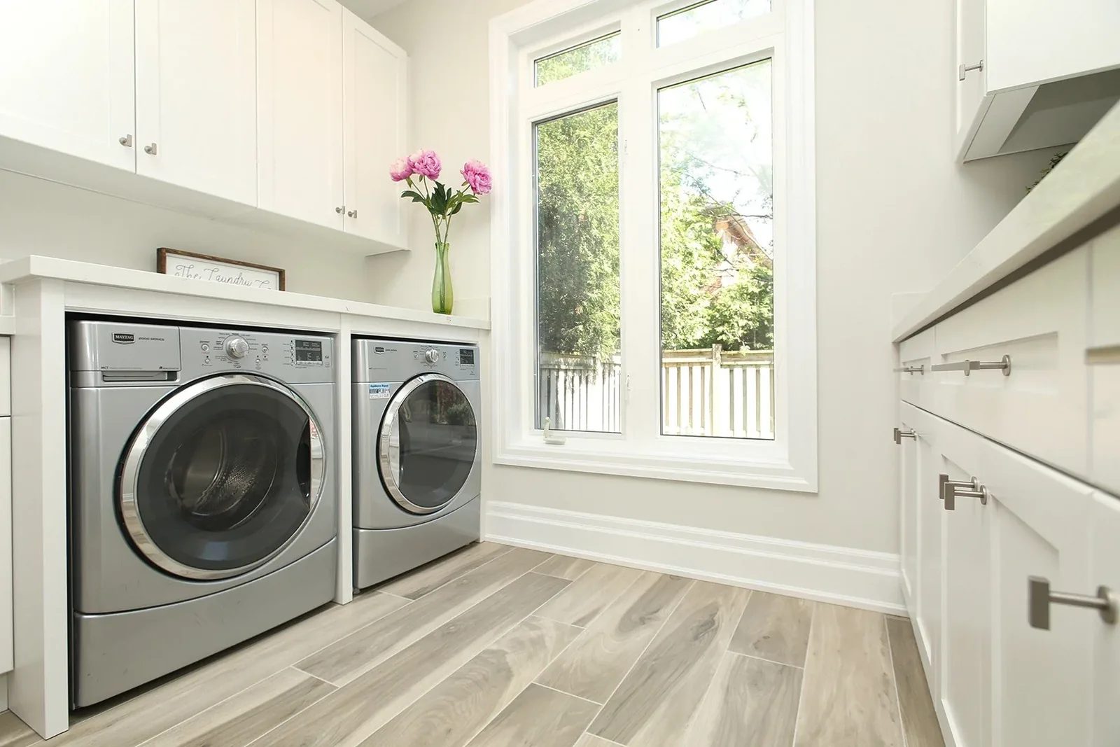 Bright laundry room with white cabinetry, silver front-load machines, and a tall window.