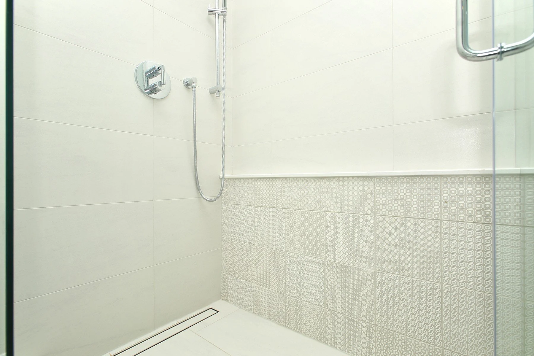 Bright walk-in shower with white tile, patterned accent walls, chrome fittings, and a linear drain.