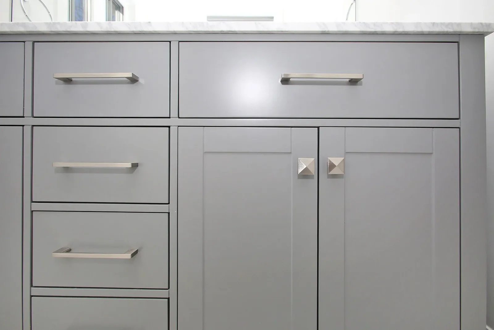 Close-up of a grey shaker bathroom vanity with brushed nickel bar pulls, pyramid knobs, and Carrara marble top.