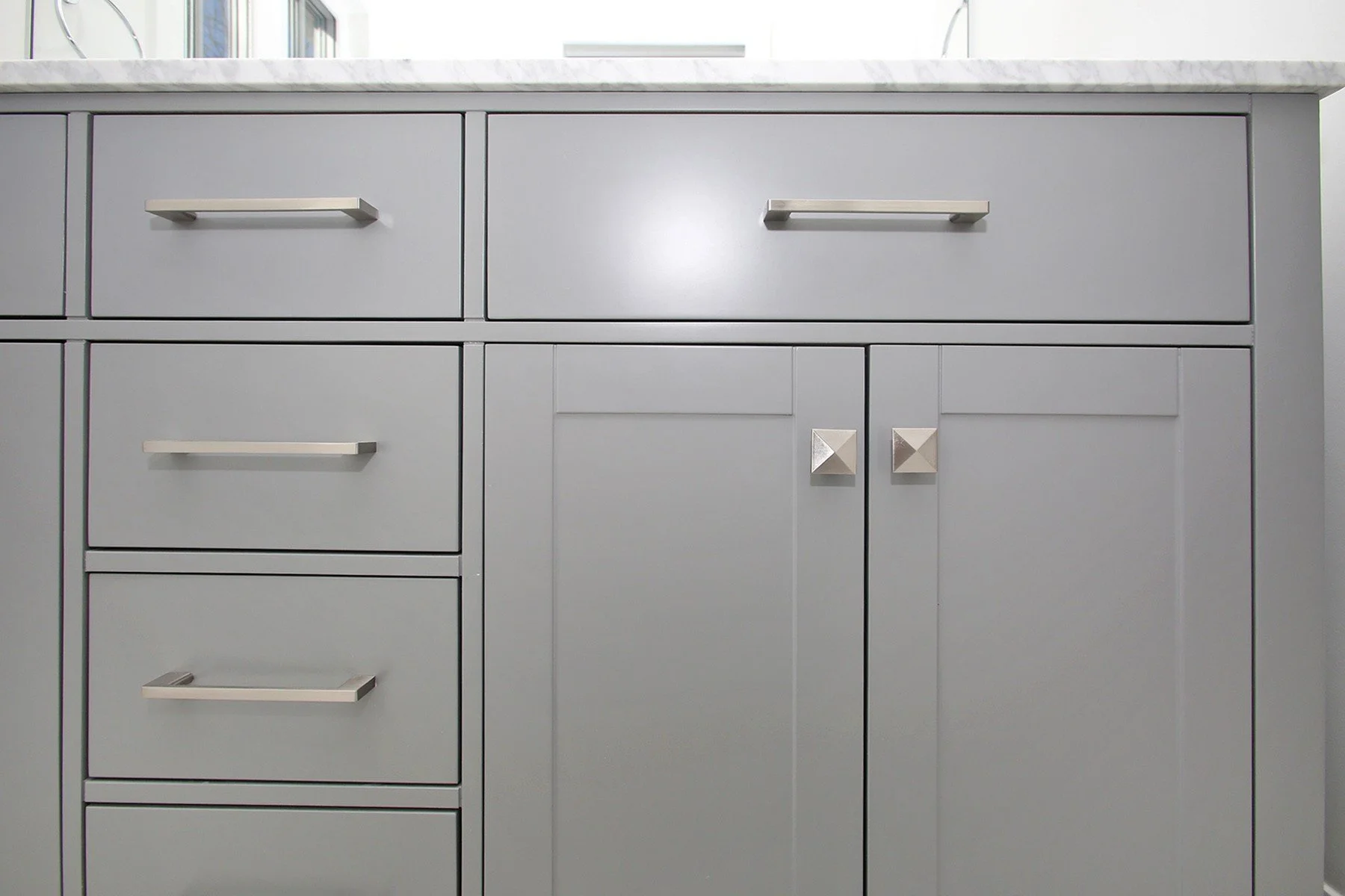 Close-up of a grey shaker bathroom vanity with brushed nickel bar pulls, pyramid knobs, and Carrara marble top.