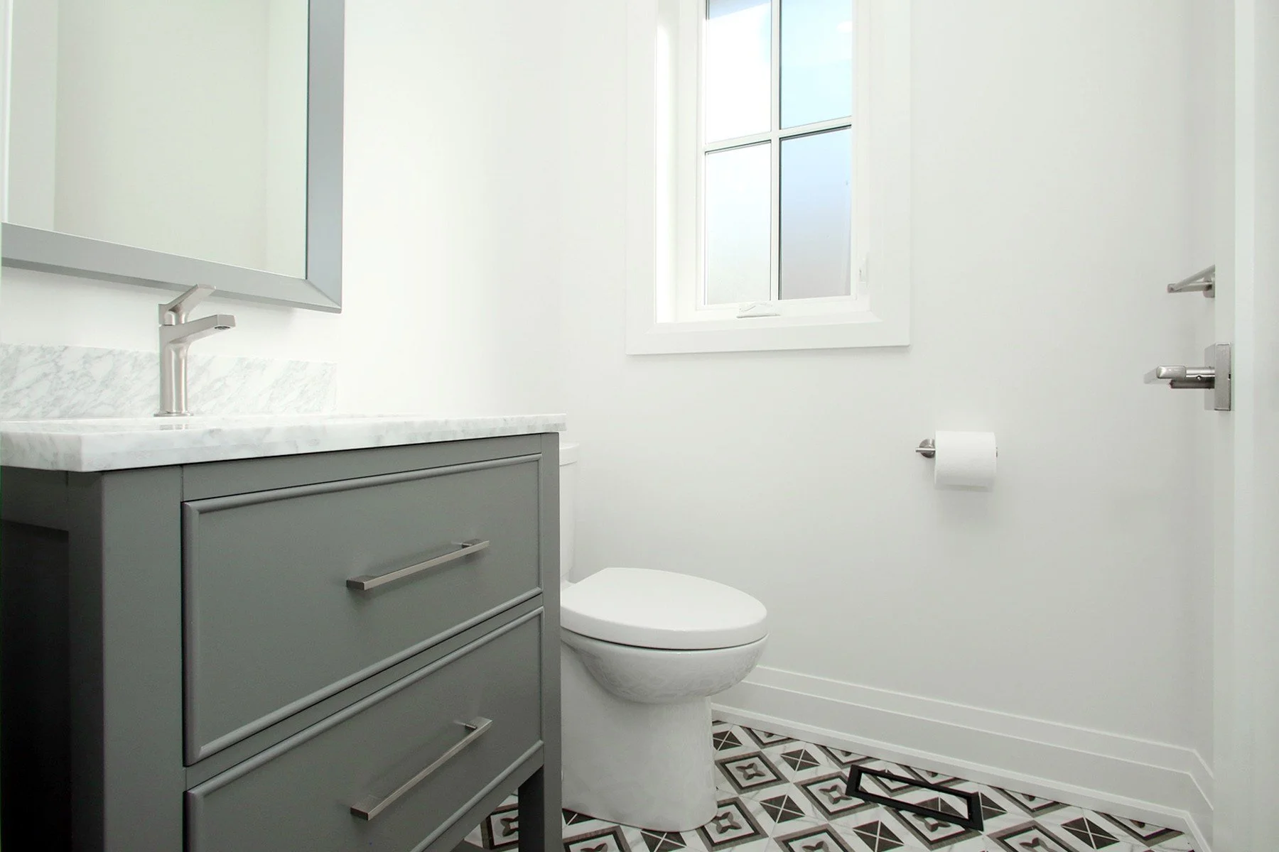 Powder room with grey single vanity, Carrara marble top, and patterned black-and-white encaustic-look floor tile.