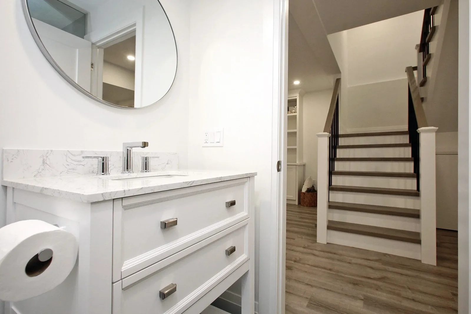 Basement bathroom white shaker vanity with round mirror, marble-look quartz top, and view of staircase beyond.