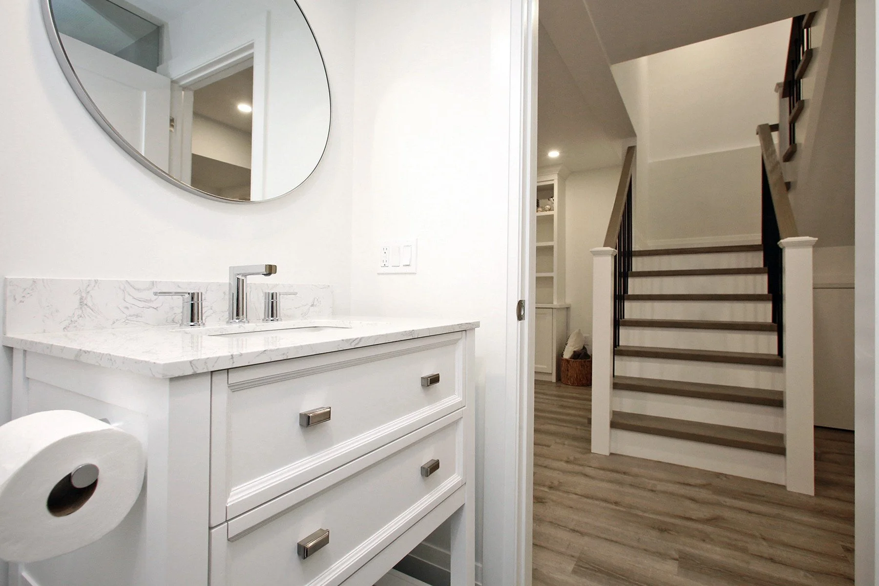 Basement bathroom white shaker vanity with round mirror, marble-look quartz top, and view of staircase beyond.