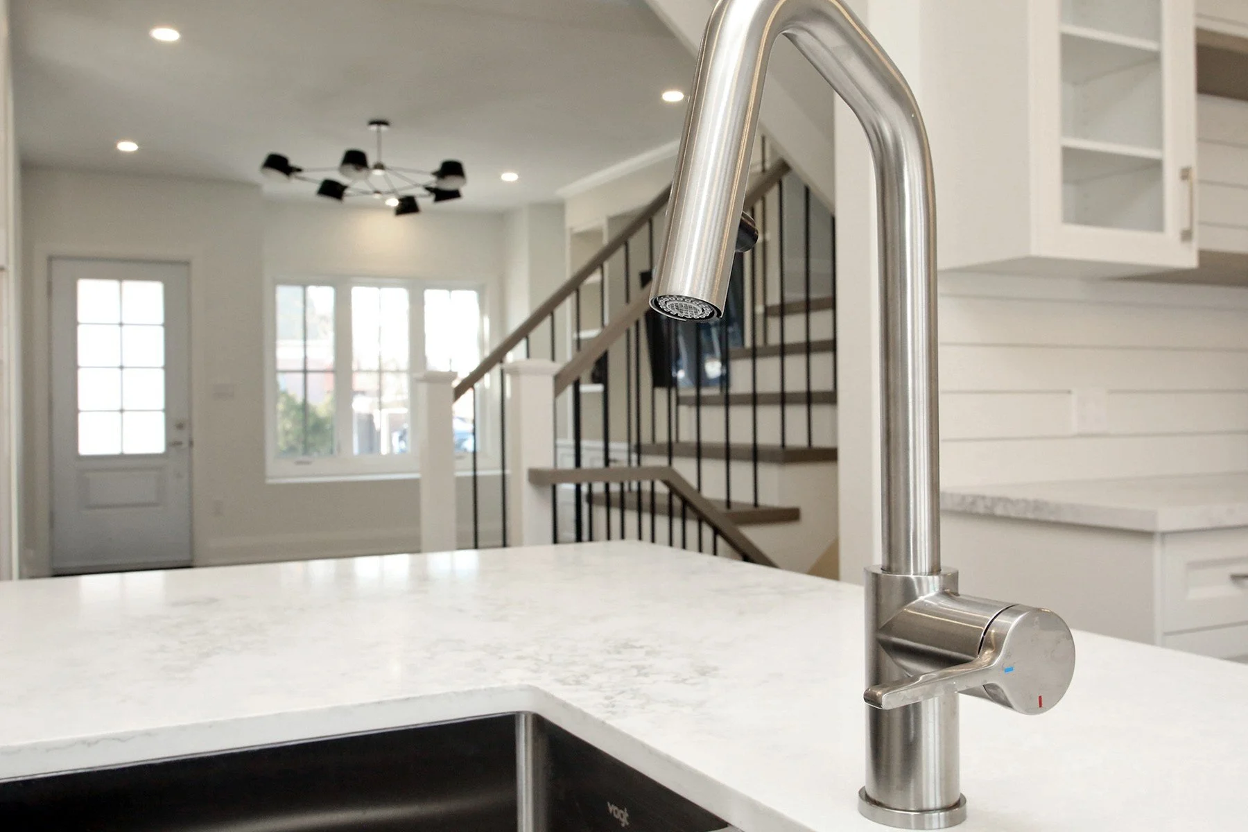 Brushed stainless gooseneck pull down kitchen faucet on a marble look quartz island with a staircase behind.