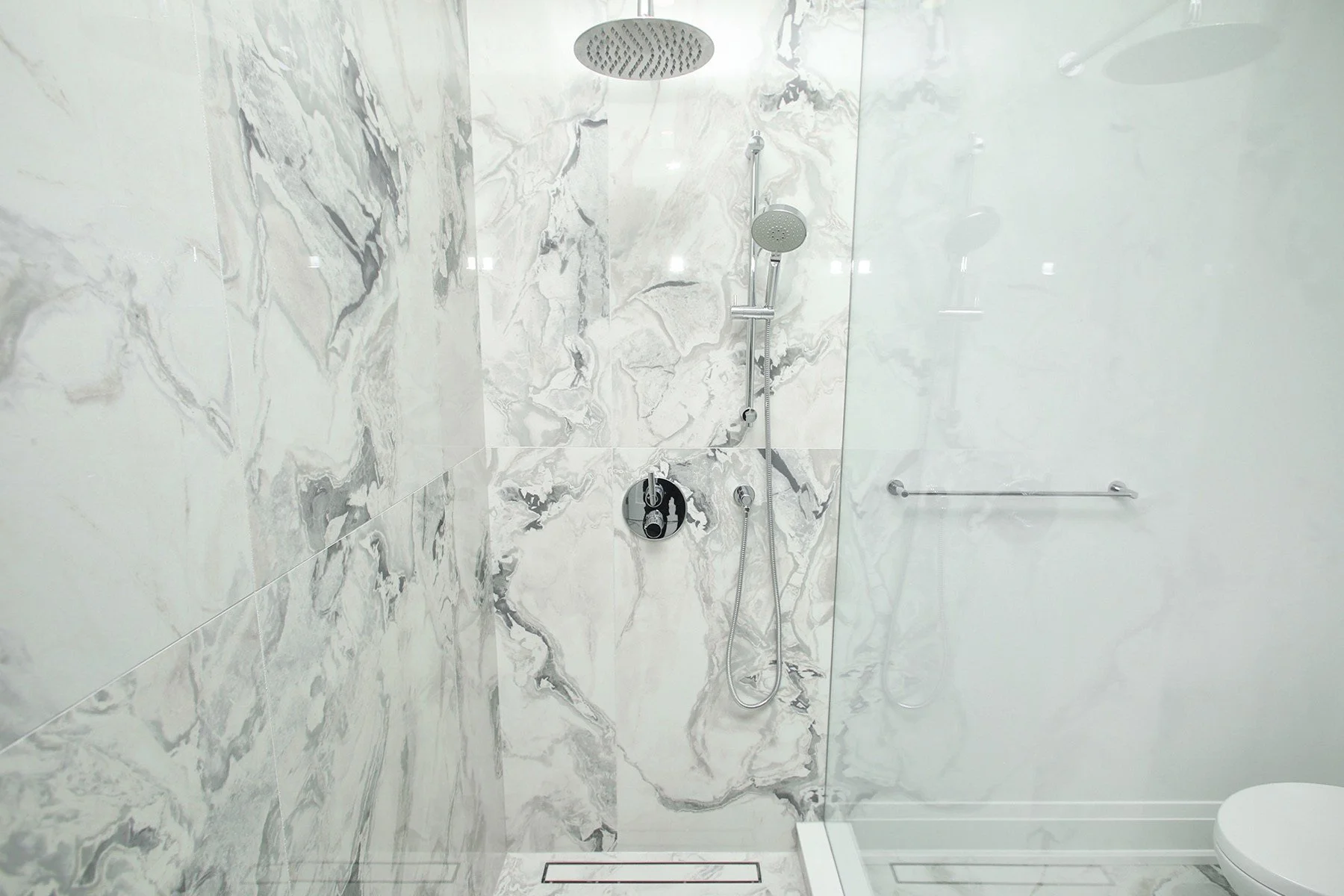 Close-up of walk-in shower with book-matched marble slab walls rain head and chrome handheld fixture.