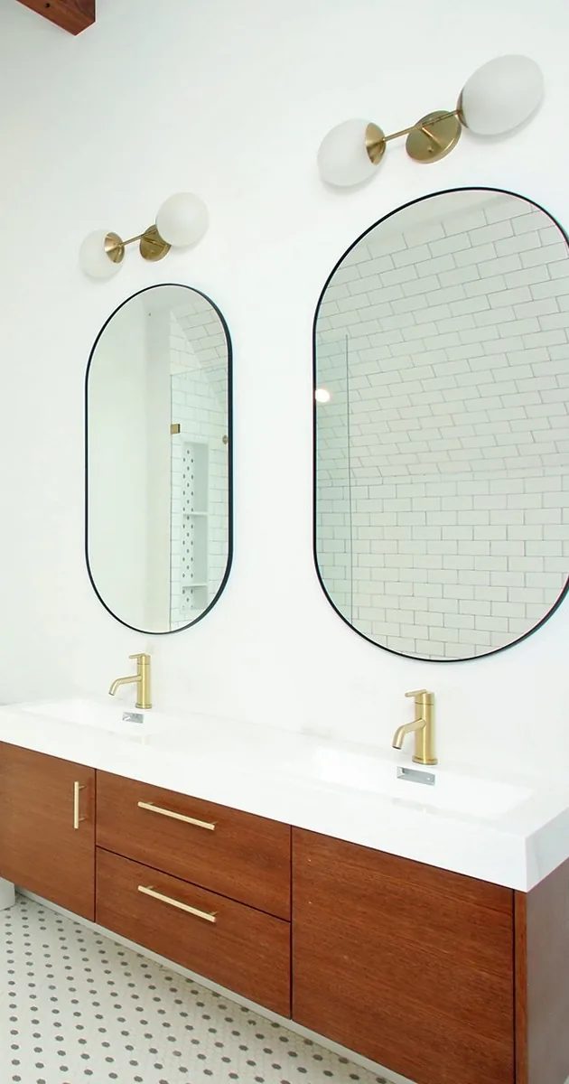Bathroom with a floating walnut double vanity, brass faucets, oval black-framed mirrors, and brass globe sconces.