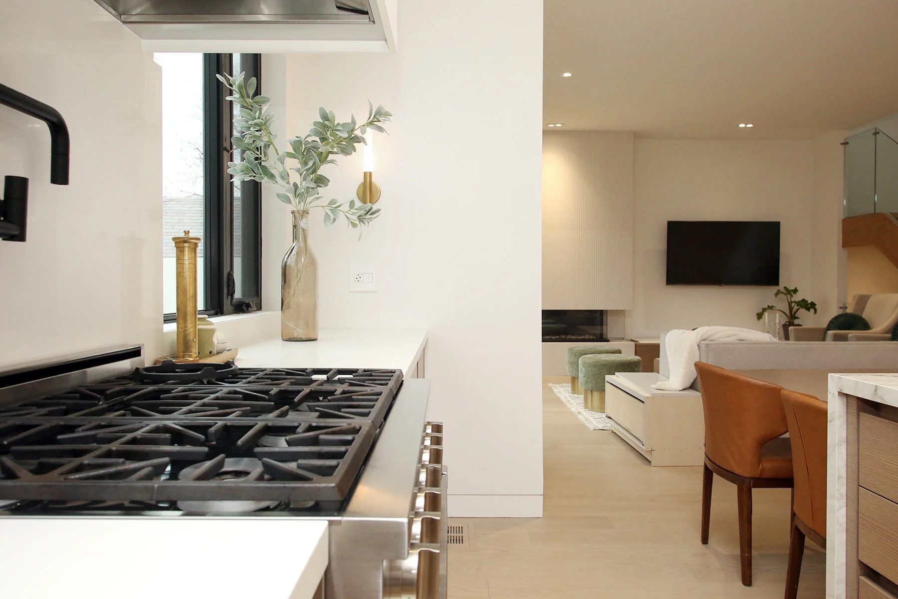 Gas cooktop in the foreground looks across an open-concept living room with a linear fireplace, wall TV, and cognac lounge chairs.