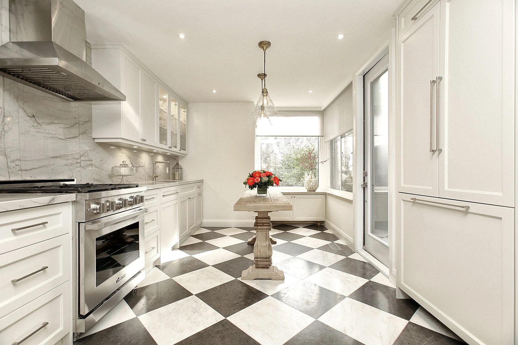 Bright white kitchen with checkered tile flooring, stone surfaces, a small centre table, and large windows.