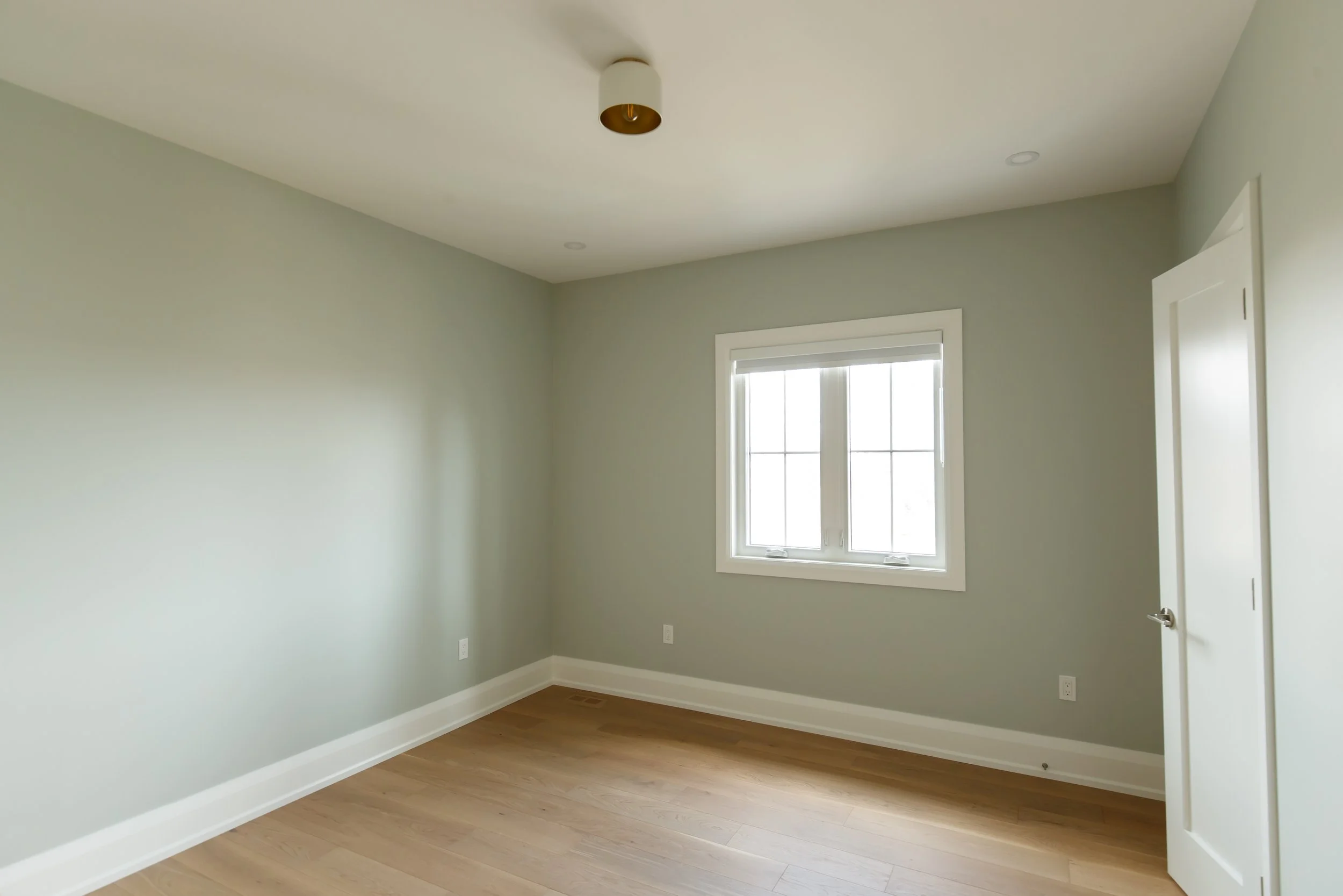 Empty sage-green bedroom with brass flush-mount ceiling light and bright casement windows.